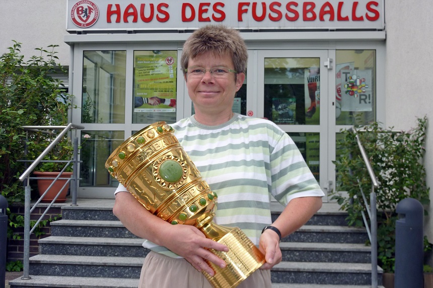 Vor dem Eingang eines Gebäudes mit der Aufschrift „HAUS DES FUSSBALLS“ steht eine Person und hält einen großen, goldfarbenen Pokal in beiden Händen. Die Person ist mittig im Bild positioniert. Der Eingang besteht aus zwei Glastüren, die von grünen Pflanzen flankiert werden. An den Türen und Fenstern sind mehrere Poster oder Schilder angebracht, deren Inhalte nicht klar erkennbar sind. Die Szene wirkt feierlich und sportlich geprägt.