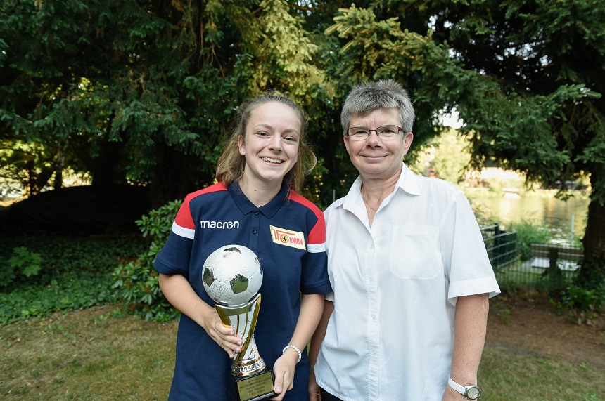 Zwei Personen stehen nebeneinander im Freien vor einer Kulisse aus grünen Bäumen und Sträuchern. Im Hintergrund sind ein Zaun und möglicherweise ein Gewässer zu erkennen. Die Person auf der linken Seite trägt ein dunkelblaues Sporttrikot mit roten und weißen Streifen auf den Schultern. Auf dem Trikot sind zwei Logos sichtbar: eines mit der Aufschrift „macron“ und ein weiteres mit „1. FC Union“. Diese Person hält einen Pokal in Form eines Fußballes in der Hand. Die Person auf der rechten Seite trägt ein weißes, kurzärmeliges Hemd. Beide Personen stehen aufrecht und blicken in Richtung Kamera.