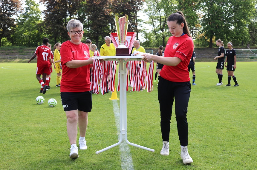 Zwei Personen in roten T-Shirts tragen gemeinsam ein Tischgestell, auf dem ein großer Pokal und zahlreiche Medaillen liegen. Sie befinden sich auf einem Fußballfeld. Im Hintergrund sind mehrere weitere Personen zu sehen – einige tragen rote Trikots, andere schwarze. Die Szene wirkt wie eine Preisverleihung oder ein sportliches Fest. Die Umgebung ist grün, mit Bäumen im Hintergrund, und vermittelt eine freundliche, gemeinschaftliche Atmosphäre.