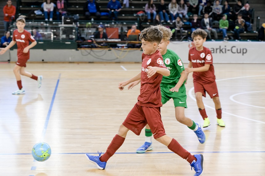 Das Foto zeigt ein Hallenfußballspiel mit jungen Spielerinnen. Die Szene spielt sich in einer Sporthalle ab. Im Vordergrund sind drei Spielerinnen zu sehen: Zwei tragen rote Trikots, eine*r ein grünes. Der Spieler oder die Spielerin im roten Trikot auf der linken Seite des Bildes führt den Ball. Der Spieler oder die Spielerin im grünen Trikot steht direkt davor und scheint zu versuchen, den Ball zu blockieren. Ein weiterer Spieler oder eine Spielerin im roten Trikot ist rechts im Hintergrund sichtbar, und eine vierte Person im roten Trikot befindet sich weiter hinten links im Bild.  Im Hintergrund sind Zuschauer*innen auf den Tribünen zu sehen. Rechts im Bild hängt ein Banner mit der Aufschrift „Sparki-Cup“, was auf ein Turnier oder eine Veranstaltung hinweist. Die Halle ist gut beleuchtet, und der Boden besteht aus hellem Hallenparkett.