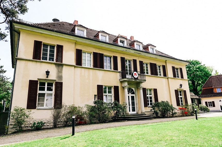 Das Foto zeigt die Vorderansicht eines großen, zweistöckigen Gebäudes, vermutlich die Sportschule Wannsee. Die Fassade ist gelb gestrichen, das Dach besteht aus braunen Ziegeln. Mehrere Fenster mit braunen Fensterläden sind sichtbar – einige davon geöffnet. In der Mitte des Gebäudes befindet sich ein Eingang mit einer weißen Tür. Direkt über dem Eingang ist ein kleiner Balkon angebracht, auf dem ein Schild mit einem roten Kreis und weißen Buchstaben hängt. Vor dem Gebäude erstreckt sich ein gepflegter grüner Rasen, und entlang der Fassade wachsen einige Büsche. Im rechten Hintergrund ist ein weiteres, kleineres Gebäude zu erkennen.