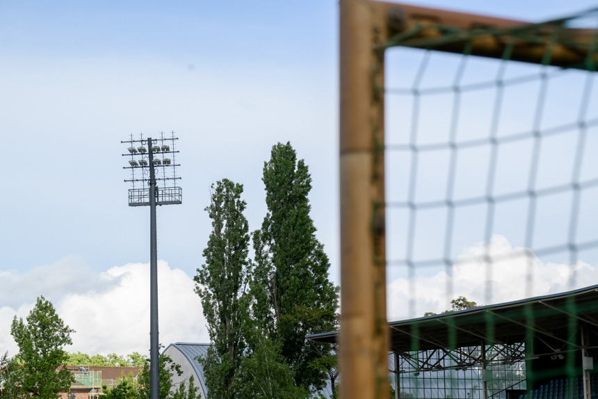 Das Foto zeigt eine Szene auf einem Fußballfeld. Rechts im Vordergrund steht ein Fußballtor mit weißem Rahmen und Netz. Der Boden ist mit grünem Rasen bedeckt. Im Hintergrund erstrecken sich hohe, grüne Bäume, die einen natürlichen Rahmen bilden. Links hinter dem Tor ragt ein hoher Flutlichtmast in den Himmel. Weiter hinten sind zwei Gebäude zu sehen: eines hat eine gewölbte Dachstruktur, das andere eine moderne Glasfassade. Der Himmel ist überwiegend blau und mit einigen weißen Wolken durchzogen. Es sind keine Menschen oder Tiere auf dem Bild zu sehen.