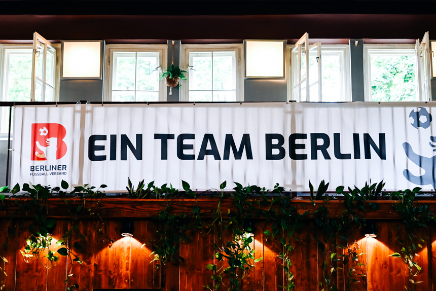 Banner mit Text „Ein Team Berlin“ hängt vor hellen Fenstern, darunter Holzwand mit vielen Pflanzen. Foto: Sandra Ritschel