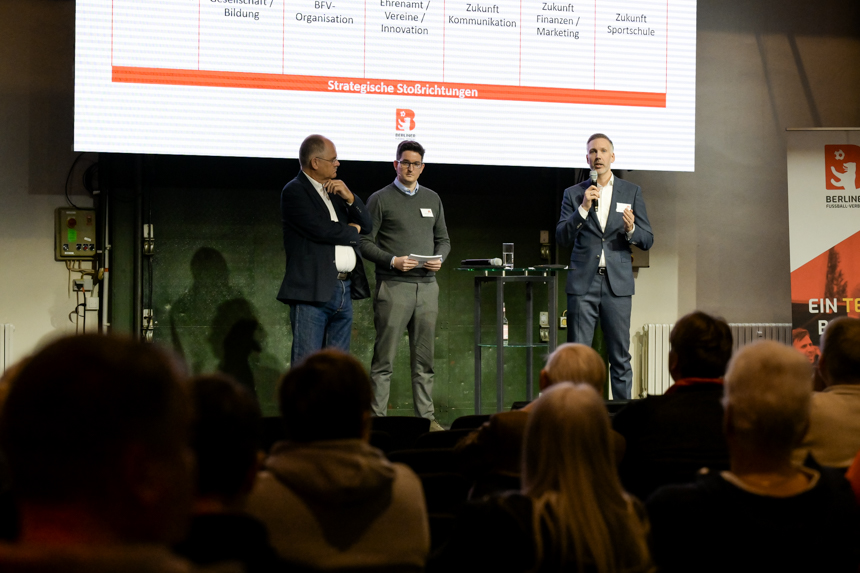 Drei Personen stehen auf einer Bühne und sprechen vor Publikum, während hinter ihnen eine Präsentationsfolie angezeigt wird. Foto: Sandra Ritschel