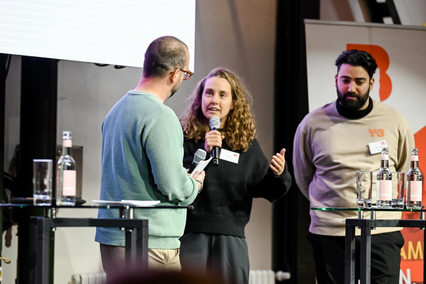 Drei Personen stehen auf einer Bühne im Gespräch, halten Mikrofone und sprechen vor einem kleinen Stehtisch mit Wasserflaschen. Foto: Sandra Ritschel