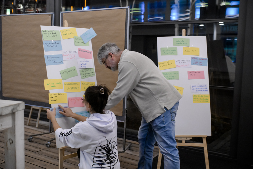 Zwei Personen gestalten eine Pinnwand und heften bunte Notizzettel an, während sie gemeinsam an Ideen und Themen arbeiten. Foto: Sandra Ritschel