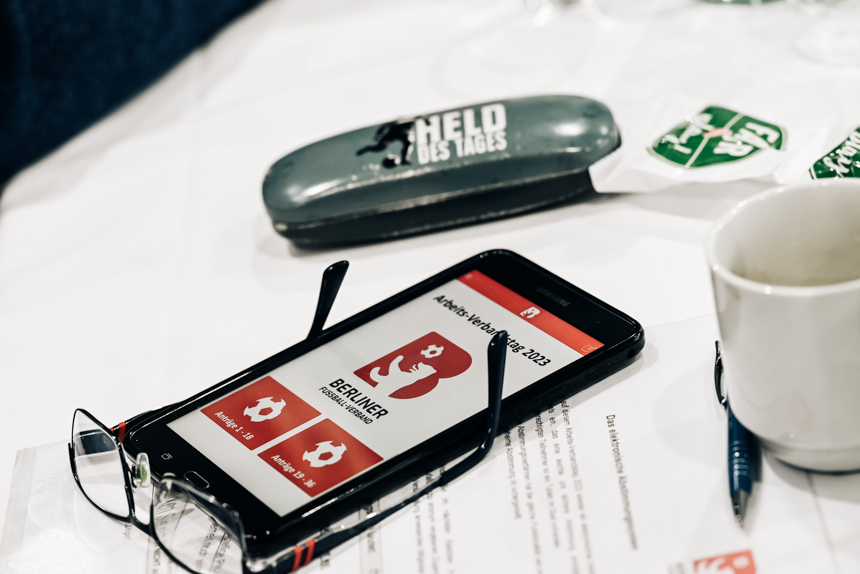 Smartphone with glasses on a table, display "Antrag Verbandstag 2023" of the Berlin Football Association, next to it cup and case "Hero of the Day". Photo: Sandra Ritschel