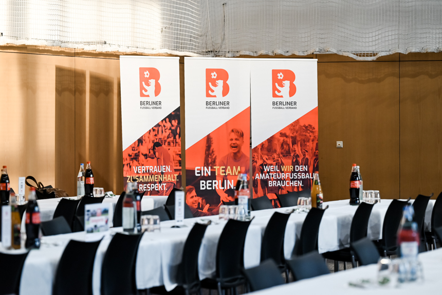 The picture shows a bright, modern function room with long, rectangular tables arranged in rows. The tables are covered with white tablecloths and equipped with bottles of drinks - mainly cola and water - as well as glasses and small table displays. Black chairs are arranged neatly at the tables. Three large, upright banners of the Berlin Football Association can be seen in the background. The banners are designed in shades of red and white and bear the association's logo with a stylized bear.