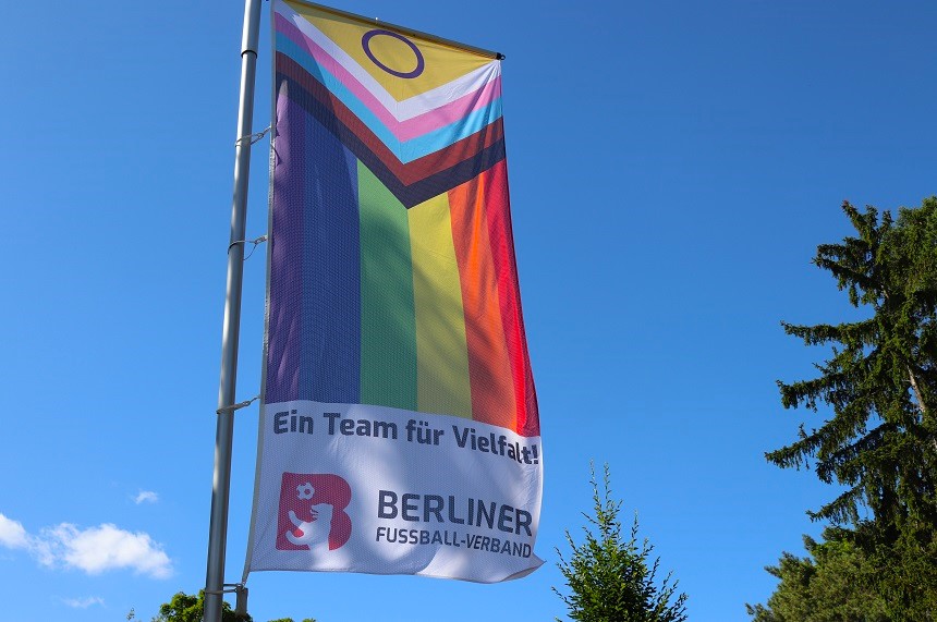 Das Foto zeigt eine Fahne, die an einem Fahnenmast im Freien hängt. Der Himmel ist klar und blau, im Hintergrund sind grüne Bäume zu sehen. Die Fahne trägt das Design der „Progress Pride Flag“, die für Vielfalt und Inklusion steht. Sie kombiniert die klassischen Regenbogenfarben mit zusätzlichen Streifen in Schwarz, Braun, Hellblau, Rosa und Weiß sowie einem gelben Kreis auf lila Hintergrund, der für Intergeschlechtlichkeit steht. Im unteren Bereich der Fahne befindet sich ein weißes Feld mit der Aufschrift: „Ein Team für Vielfalt!“ Darunter ist das Logo des Berliner Fußball-Verbandes (BFV) zu sehen, bestehend aus einem roten Löwen und dem Schriftzug „BERLINER FUSSBALL-VERBAND“ in Schwarz. Die Fahne vermittelt eine klare Botschaft für Akzeptanz, Respekt und Zusammenhalt im Sport und darüber hinaus.