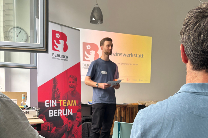 A person stands in front of a group and speaks, holding cue cards; banners reading “Berlin Football Association” and “Club Workshop”; participants in the foreground. Photo: BFV
