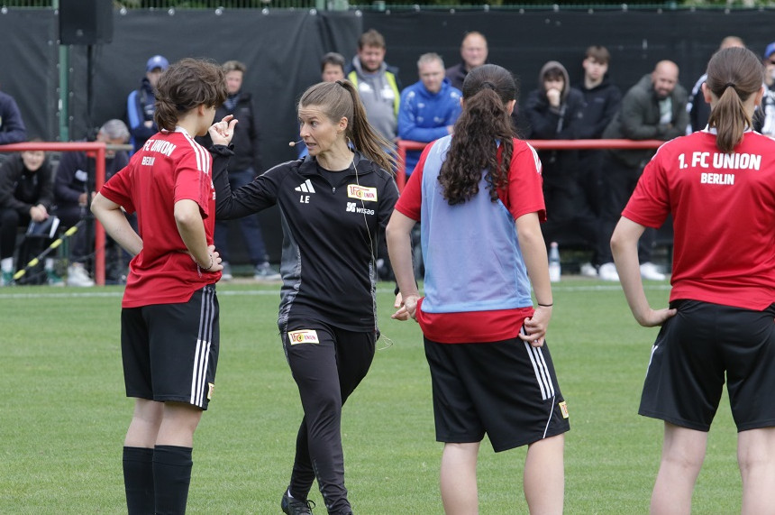 Auf dem Foto ist eine Szene auf einem Fußballfeld zu sehen. Im Vordergrund stehen vier Personen. Drei von ihnen tragen rote Trikots mit der Aufschrift „1. FC UNION BERLIN“ auf dem Rücken. Die vierte Person trägt schwarze Sportkleidung und scheint Anweisungen zu geben – möglicherweise handelt es sich um eine Trainerin oder einen Trainer. Die Personen stehen in einem Halbkreis und wirken konzentriert. Im Hintergrund sind weitere Menschen zu erkennen, die das Geschehen beobachten. Der Rasen ist grün und gut gepflegt, und das Wetter scheint freundlich zu sein.
