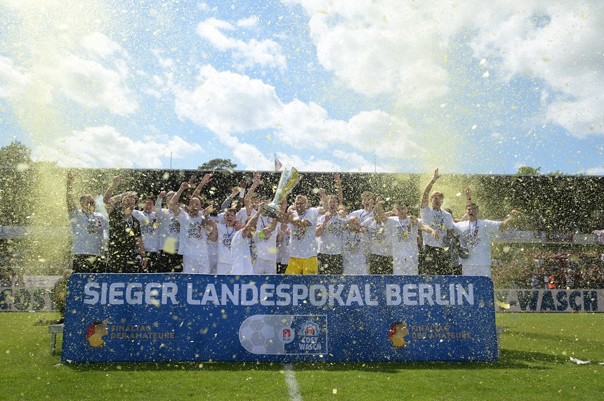 Das Foto zeigt eine jubelnde Fußballmannschaft, die den Gewinn des Landespokals Berlin feiert. Die Spieler tragen weiße Trikots und stehen hinter einem auffälligen blauen Banner mit der Aufschrift „SIEGER LANDESPOKAL BERLIN“. Einige Teammitglieder halten stolz den Pokal in die Höhe, während andere mit erhobenen Armen den Moment feiern. Im Hintergrund erkennt man Zuschauer auf einer Tribüne, die das Geschehen verfolgen. Zusätzlich verstärken versprühter Champagner oder Konfetti die festliche und ausgelassene Stimmung des Augenblicks.