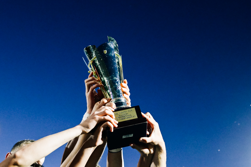 Mehrere Hände unterschiedlicher Hauttöne halten gemeinsam einen silbernen Pokal in die Höhe. Der Pokal ist glänzend, hat zwei seitliche Griffe und steht auf einer breiten Basis. Auf dem Sockel befindet sich eine goldene Plakette mit einer Inschrift, die auf dem Bild nicht lesbar ist. Der Hintergrund zeigt einen wolkenlosen, klaren blauen Himmel. Die Szene vermittelt ein Gefühl von Gemeinschaft, Zusammenarbeit und gemeinsamer Leistung, ohne dass einzelne Personen im Vordergrund stehen.