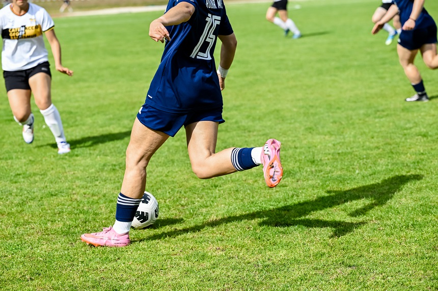 A soccer scene is taking place on a green grass field. In the center of the frame is a person wearing dark blue athletic clothing with the number 15 on the back. The person is dribbling a black-and-white soccer ball and is wearing pink soccer cleats and dark blue socks with white stripes. Only part of their upper body and head are visible in the frame.  To the left, right, and in the background are other people in athletic wear who are also moving around on the field. One person on the left is wearing a white jersey with a black and yellow stripe across the chest. The people in the background are blurred due to the shallow depth of field.  The scene was captured in bright daylight. The grass glows a vibrant green, and the deep shadows on the ground indicate sunny weather. The focus is on the movement of the game and the central figure’s possession of the ball. Overall, the photo conveys a dynamic moment during a soccer game.