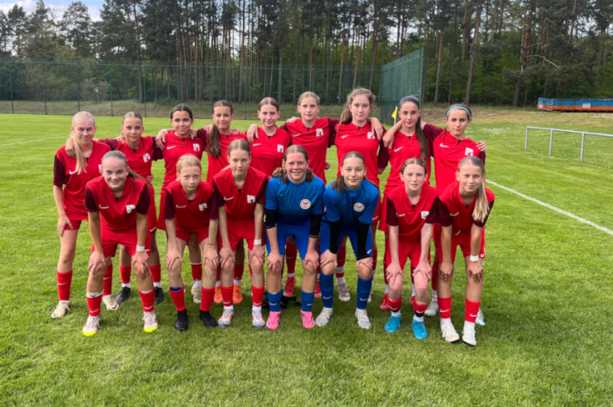 Das Foto zeigt eine Gruppe von 15 Personen, die in zwei Reihen auf einem Fußballfeld posieren. Die vordere Reihe besteht aus acht Personen, die in roten Trikots und schwarzen Shorts gekleidet sind. Zwei Personen in der Mitte tragen blaue Trikots und Shorts. Die hintere Reihe besteht aus sieben Personen, die ebenfalls rote Trikots und schwarze Shorts tragen. Alle stehen oder hocken auf grünem Gras mit einem Wald im Hintergrund.
