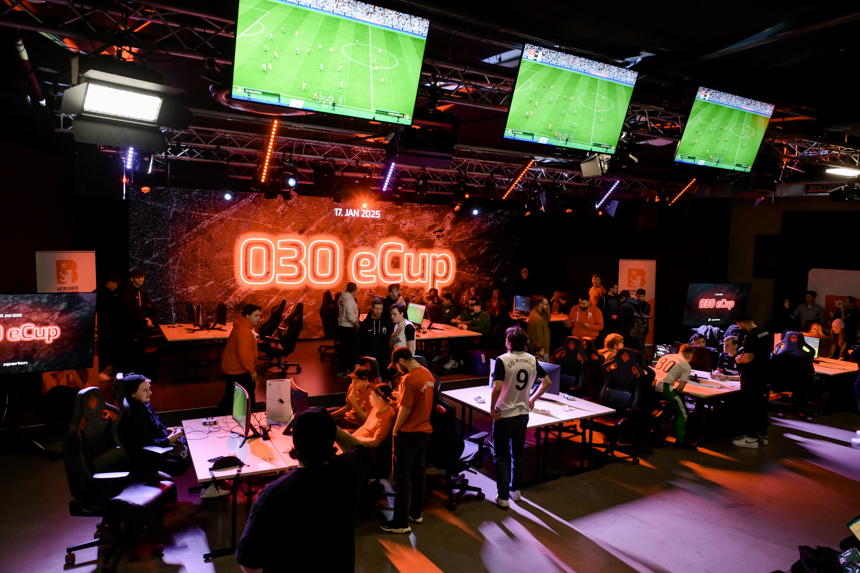  Event room of the 030 eCup with several gaming stations. Participants sit at round tables with computers and play FIFA. Three large screens above the scene show games in progress. An orange illuminated stage with the "030 eCup" logo is visible in the background.