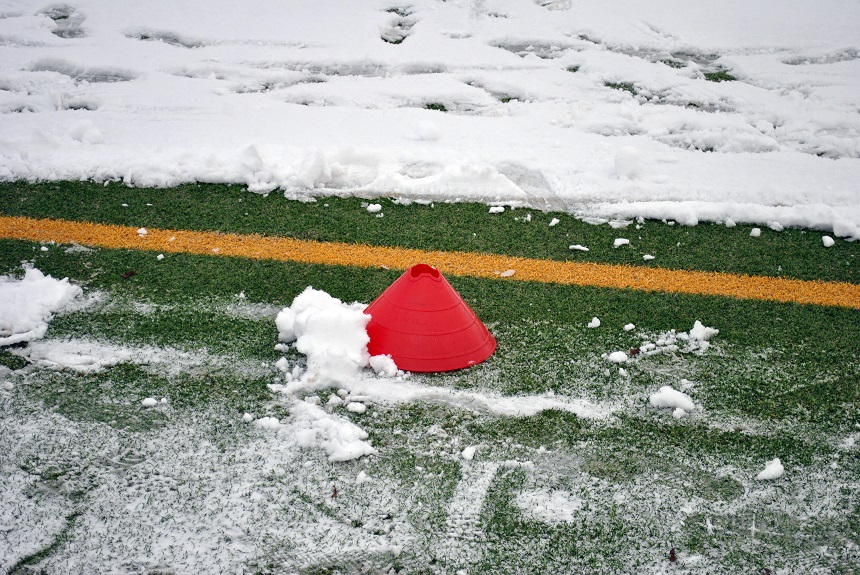 Das Bild zeigt einen roten Markierungshütchen auf einem Kunstrasenplatz. Der Boden ist teilweise mit Schnee bedeckt, wobei einige Bereiche des grünen Rasens sichtbar bleiben. Quer durch das Bild verläuft eine gelbe Linie, die vermutlich eine Spielfeldmarkierung darstellt. Rund um das Hütchen liegen kleine Schneehäufchen, und im Hintergrund sind Fußspuren im Schnee erkennbar. Die Szene vermittelt den Eindruck eines Sportplatzes im Winter, auf dem kürzlich Schnee gefallen ist.