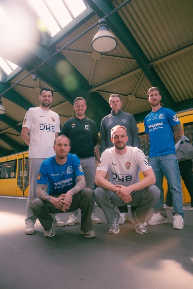  Six people in soccer shirts and leisure wear pose under a station roof in front of a yellow subway train. Two are squatting in front, four are standing behind them.