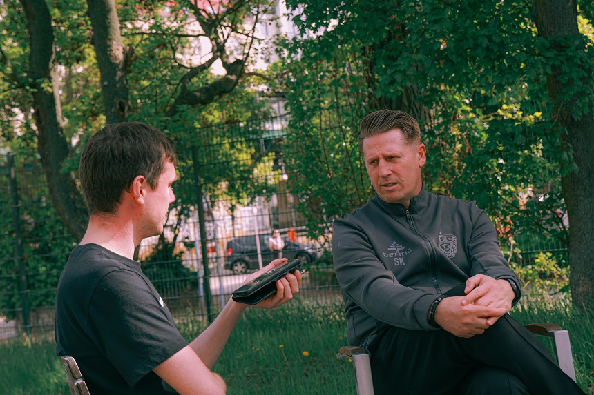 Two people are sitting outside talking. The person on the left holds a smartphone or recording device in their hand and points it at the other person, who is relaxed and listening with their hands clasped.