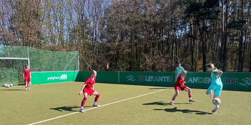 Fußballspielsituation auf einem Kunstrasenplatz. Mehrere Jugendspieler:innen tragen rote und türkise Trikots. Eine Person in türkisem Trikot führt den Ball, während zwei Gegenspieler:innen in roten Trikots verteidigen. Im Hintergrund sind ein Tor, eine grüne Bande mit der Aufschrift „Tolerantes Brandenburg“ sowie Bäume entlang des Spielfeldrands zu sehen.