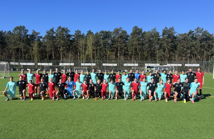 Gruppenfoto mehrerer Jugendfußballmannschaften auf einem grünen Kunstrasenplatz. Rund 40 Personen stehen und knien in einer Reihe und tragen unterschiedliche Trikots in Rot, Türkis, Schwarz und Blau. Im Hintergrund sind Fußballtore, ein Zaun, parkende Autos sowie ein dichter Wald unter blauem Himmel zu sehen.