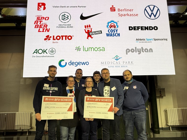 Eine Gruppe von Menschen steht gemeinsam auf einer Bühne vor einer großen Leinwand, auf der die Logos zahlreicher Partnerorganisationen und Sponsoren des Berliner Fußball-Verbandes zu sehen sind. In der vorderen Reihe halten mehrere Personen zwei große symbolische Schecks des BFV, jeweils mit einem Preisgeld von 1.000 Euro. Die Personen tragen sportliche oder casual Kleidung, einige mit Vereinslogos auf Pullovern oder Jacken. Der Raum wirkt wie ein Veranstaltungs- oder Saalbereich mit professioneller Beleuchtung. Die Szene vermittelt den Moment einer feierlichen Ehrung oder Preisverleihung.