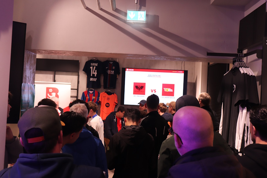 Gruppe von etwa 20 Personen in einem Laden mit Fußballartikeln (T-Shirts, Jacken) versammelt sich vor Bildschirm mit „ALBANIEN vs.“-Anzeige. Fokus auf Rückenansichten und Profile, beleuchteter Innenraum mit Ausgangshinweisen oben. Ideal für Event-Promo im Berliner Fußball-Verband-Kontext.