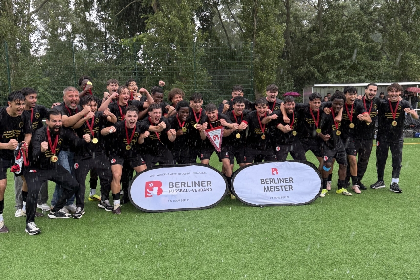 A youth soccer team in black jerseys, wearing medals and holding a championship pennant, on the soccer field in the rain after winning the Berlin championship.