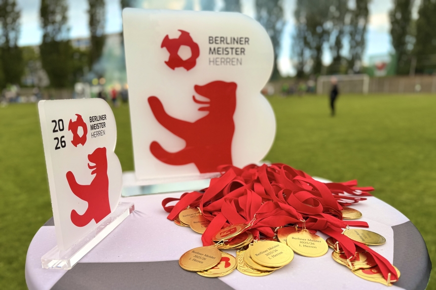 Medals and championship trophies from the Berlin Soccer Association on a table at the edge of the field; a soccer field in the background.