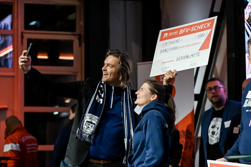 Zwei Personen stehen nebeneinander in einem Innenraum und machen gemeinsam ein Selfie. Eine der Personen hält ein Smartphone nach oben gerichtet, während die andere dicht danebensteht. Beide tragen dunkle Jacken oder Hoodies; eine Person hat zudem einen Fanschal um den Hals. Über ihnen hält eine der Personen einen großen symbolischen Scheck des Berliner Fußball-Verbandes hoch, auf dem ein Preisgeld von 1.000 Euro vermerkt ist. Im Hintergrund befinden sich weitere Personen sowie Roll-Ups und Lichtelemente, die eine Veranstaltungsszenerie andeuten. Die Atmosphäre wirkt lebhaft und festlich, passend zu einer Preisverleihung oder Ehrung.