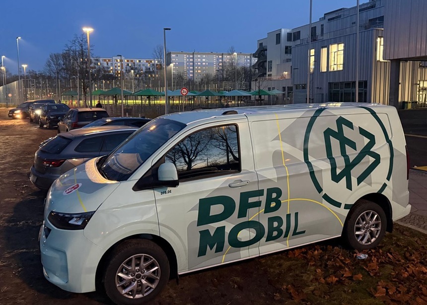 Ein helles Lieferfahrzeug mit DFB‑Mobil‑Aufdruck steht in der Dämmerung neben Autos vor modernen Gebäuden.
