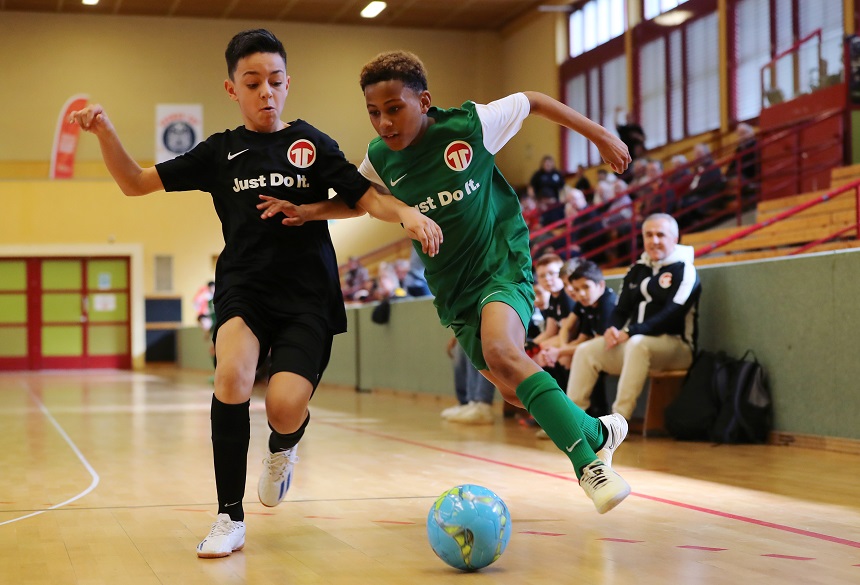 In einer Turnhalle kämpfen zwei junge Fußballspieler dynamisch um den Ball. Einer trägt ein schwarzes Trikot, der andere ein grünes. Beide befinden sich in schneller Bewegung: Der Spieler im grünen Trikot dribbelt den blau‑grünen Ball knapp über den Hallenboden, während der Spieler im schwarzen Trikot dicht neben ihm läuft und versucht, den Ball zu erreichen. Im Hintergrund sitzen Zuschauerinnen und Zuschauer auf einer Tribüne und verfolgen das Spiel. Die Halle ist hell beleuchtet, mit Holzboden, hohen Fenstern und einer sportlichen Atmosphäre.