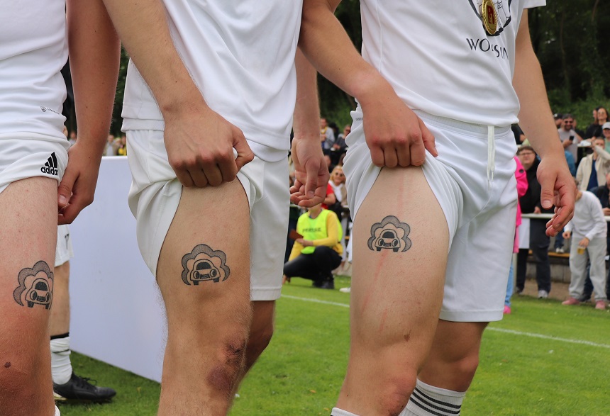 Drei Personen in weißen Sportoutfits heben ihre Shorts an und zeigen identische Tattoos auf dem Oberschenkel. Im Hintergrund stehen Zuschauer am Rand eines Sportplatzes.