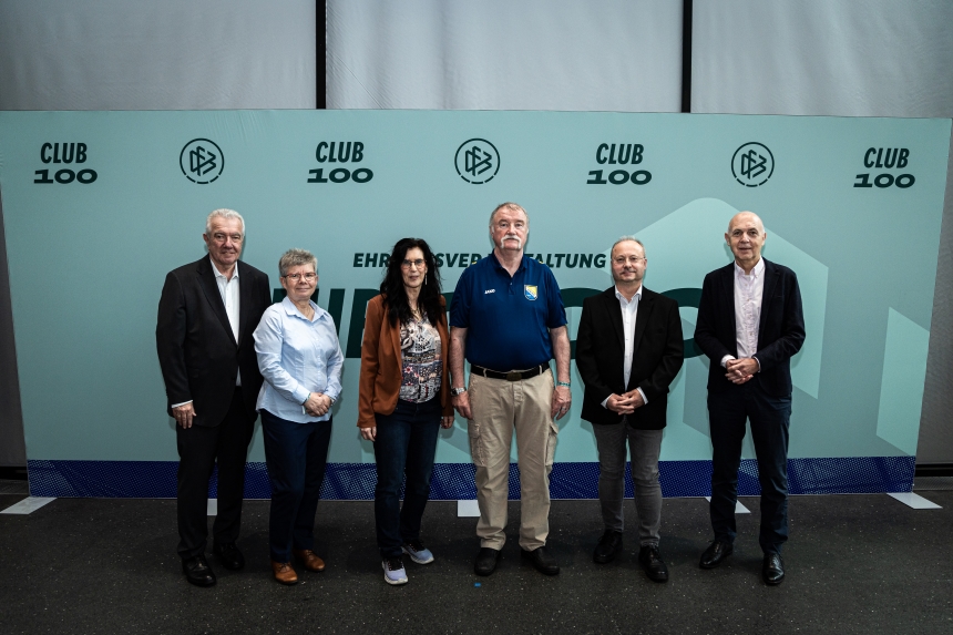 Sieben Personen stehen nebeneinander vor einer türkisen Club‑100‑Fotowand und posieren in unterschiedlicher Kleidung für ein Gruppenfoto.