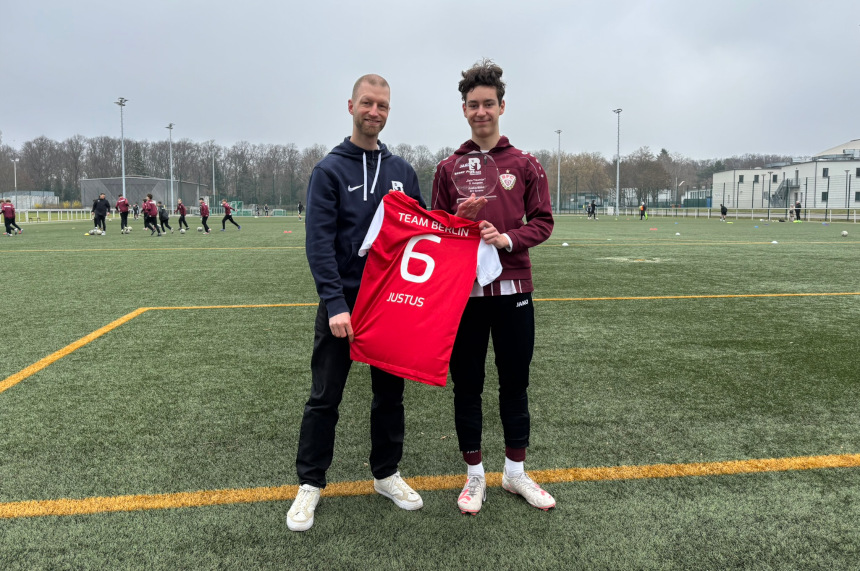 Zwei Personen stehen auf einem Fußballfeld und halten gemeinsam ein rotes Trikot mit der Rückennummer 6 und dem Namen „Justus“ hoch. Eine der Personen hat außerdem eine gläserne Auszeichnung in der Hand. Im Hintergrund sind weitere Spieler beim Training sowie Sportanlagen und umliegende Gebäude zu sehen. 
