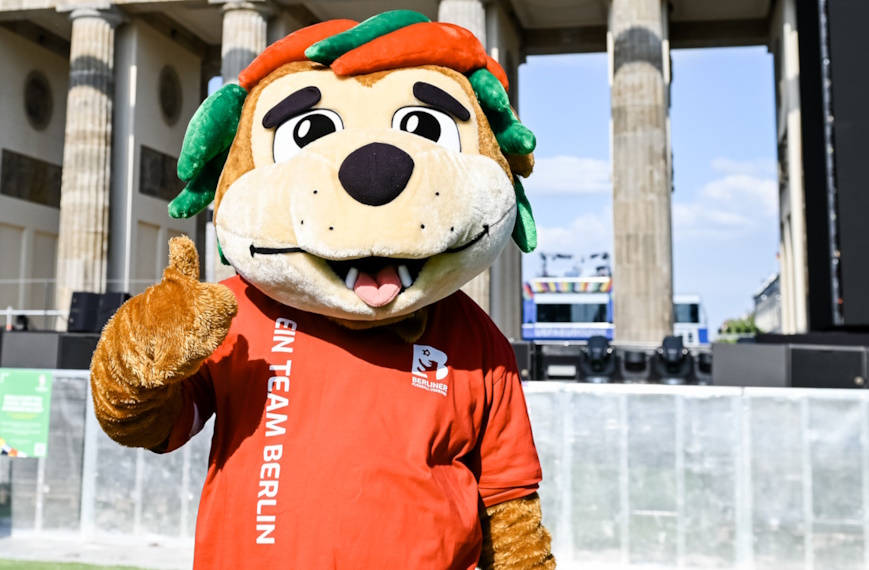 Ein fröhliches Maskottchen im Tierkostüm steht vor dem Brandenburger Tor und zeigt mit einem Daumen nach oben in die Kamera. Es trägt ein rotes T‑Shirt mit der Aufschrift „Ein Team Berlin“ und wirkt einladend und gut gelaunt.