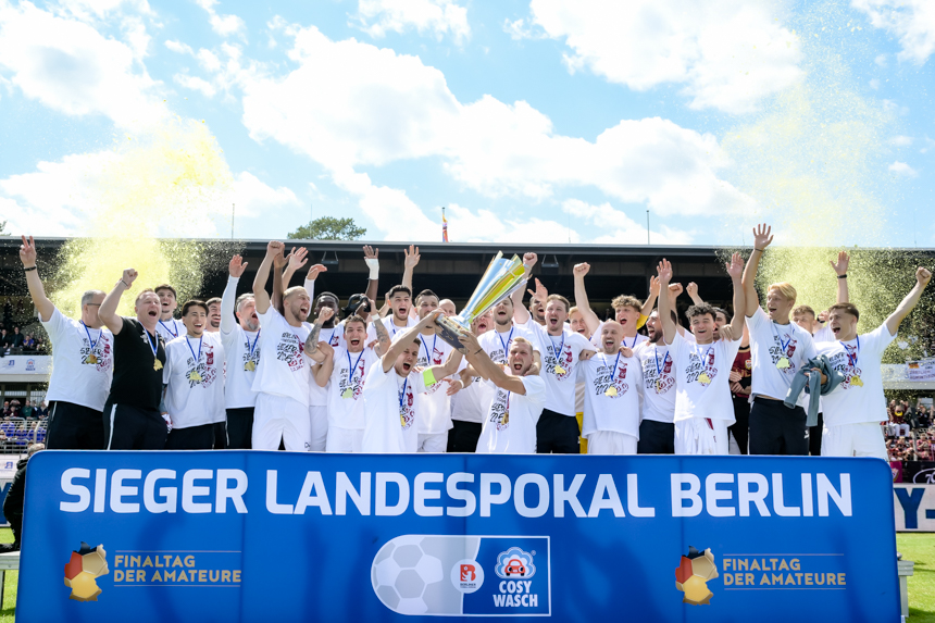 A team in white jerseys lifts a trophy together and stands cheering behind a "Winner Berlin State Cup" banner in the stadium. Photo: Sandra Ritschel