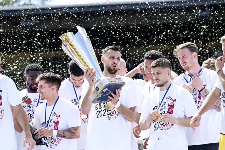 Several people in white jerseys stand close together in the stadium, one person holds a large trophy, confetti falls in the background.