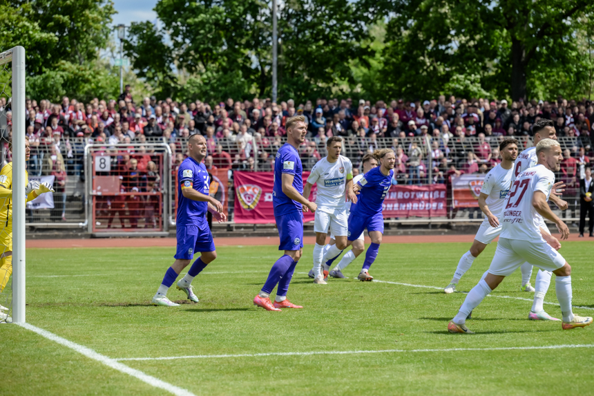 Several players in blue and white jerseys stand in the penalty area, spectators fill the stands behind the goal. Photo: Sandra Ritschel