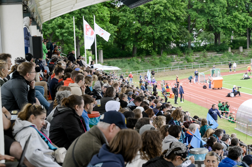 Numerous spectators sit tightly packed in the covered stands of a stadium. The stands run diagonally from left to right and are almost completely full. People in jackets and weatherproof clothing can be seen in the foreground. On the right, the view opens up onto the running track and the stadium pitch. Several people and technical equipment are standing along the track. Green trees and several waving flags are visible in the background. The photo shows a lively stadium atmosphere during a sporting event.