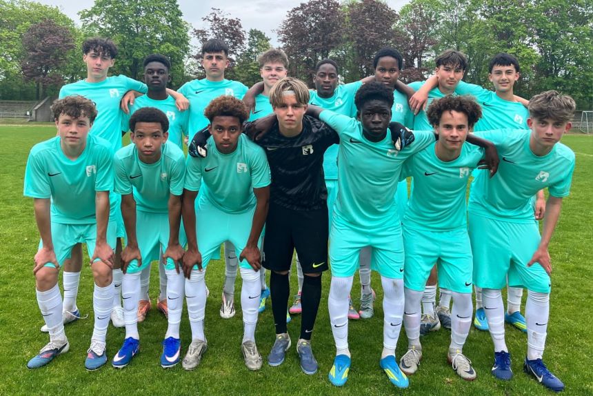 Jugend‑Fußballteam in türkisfarbenen Trikots posiert auf Rasenplatz, vorne gebeugt, hinten stehend, mit Bäumen im Hintergrund.
