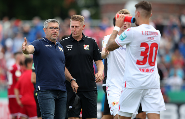 Two supervisors or coaches are talking to players from one team on the sidelines. In the foreground is a player in a white jersey with shirt number 30, while other players and a spectator stand can be seen in the background.