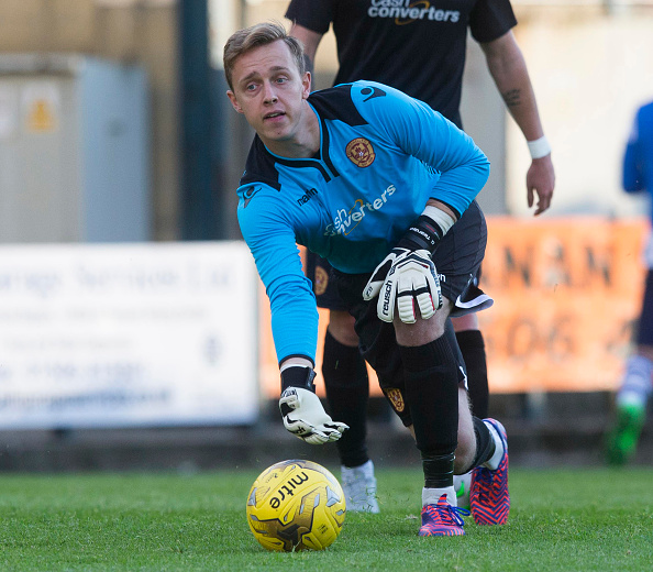 A goalkeeper in a blue jersey leans forward and grabs a yellow soccer ball on the pitch, while another player can be seen in the background.