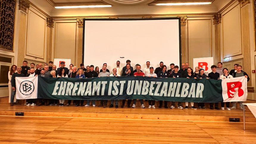 Große Gruppe in einer Halle hält ein langes Banner mit dem Text „Ehrenamt ist unbezahlbar“ vor einer großen Leinwand.