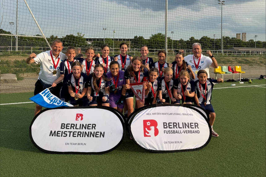 A sports team in blue-and-white jerseys, wearing medals and carrying a championship pennant, on the soccer field in front of signs for the Berlin Championship.