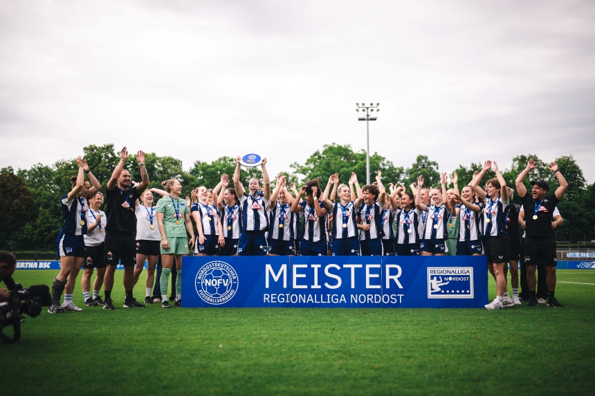 A sports team in blue-and-white jerseys, wearing medals, on a soccer field behind a sign for the Northeast Regional League championship.