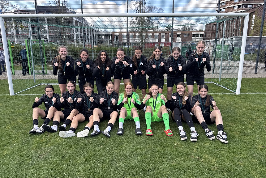 Vor einem Fußballtor posiert eine komplette Mädchenmannschaft geschlossen und gut gelaunt. Die Spielerinnen tragen schwarze Trainingsjacken, zwei in der Mitte sitzen in leuchtend grünen Torwarttrikots. Alle haben die Fäuste motiviert nach vorne gestreckt, als würden sie gemeinsam jubeln oder Teamgeist zeigen. Die Gruppe sitzt und steht auf einem gepflegten Rasenplatz, im Hintergrund sind ein Zaun, Trainingsfelder und einige Gebäude zu sehen. Die Stimmung wirkt sportlich, stolz und voller Zusammenhalt.