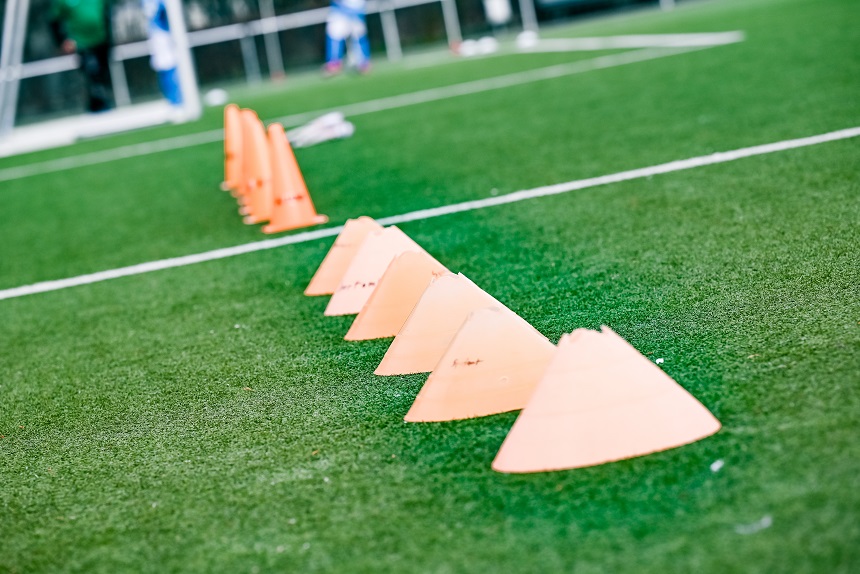 Auf einem grünen Kunstrasenplatz sind mehrere orangefarbene Markierungshütchen in einer geraden Linie aufgestellt, vermutlich zur Vorbereitung eines Fußballtrainings oder einer Übungseinheit. Im Hintergrund sind unscharf weitere Spielfeldmarkierungen, ein Tor und einzelne Personen zu erkennen.