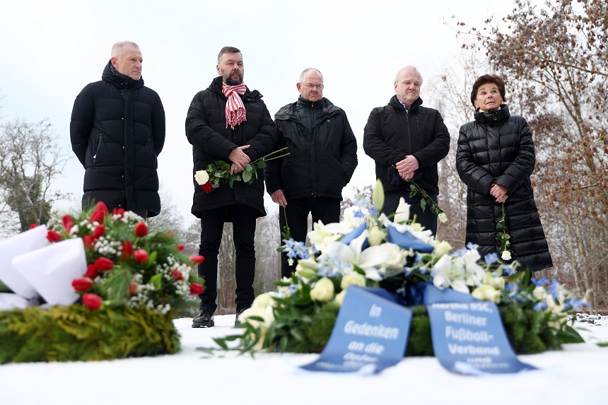 Fünf Personen stehen im Freien auf einer schneebedeckten Fläche. Sie tragen winterliche, dunkle Kleidung und halten jeweils einzelne Blumen in den Händen, überwiegend weiße Rosen. Vor ihnen liegen mehrere große Kränze und Blumenarrangements auf dem Boden. Ein Kranz besteht aus weißen und blauen Blumen und trägt zwei blaue Schleifen mit Aufschriften. Ein weiterer Kranz ist mit roten Tulpen und Tannenzweigen gestaltet. Die Szene wirkt ruhig und erinnert an eine Gedenk- oder Trauerzeremonie. Im Hintergrund sind kahle Bäume und ein winterlicher Himmel zu sehen.