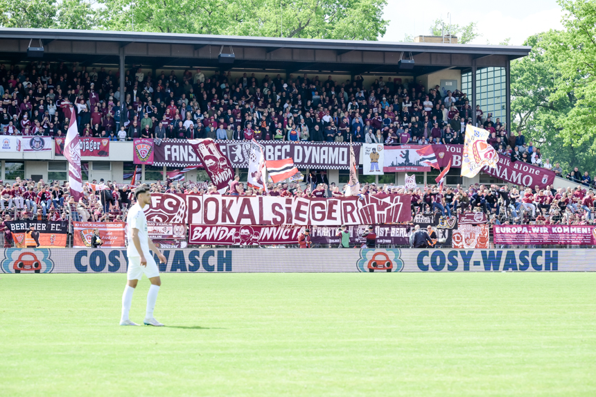 The photo shows a packed stand in a soccer stadium. Many people are holding up red and white banners and flags, including large lettering such as "POKALSIEGER". In front of the stands, a single person in a white kit is standing on the green pitch. A digital advertising banner with the words "COSY-WASCH" can be seen on the edge of the pitch.